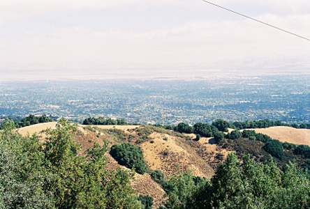 [View of a mound just below the one on which the camera is located and looking at the valley below with many trees interspersed with the buildings. The sky is very hazy in the distance.]